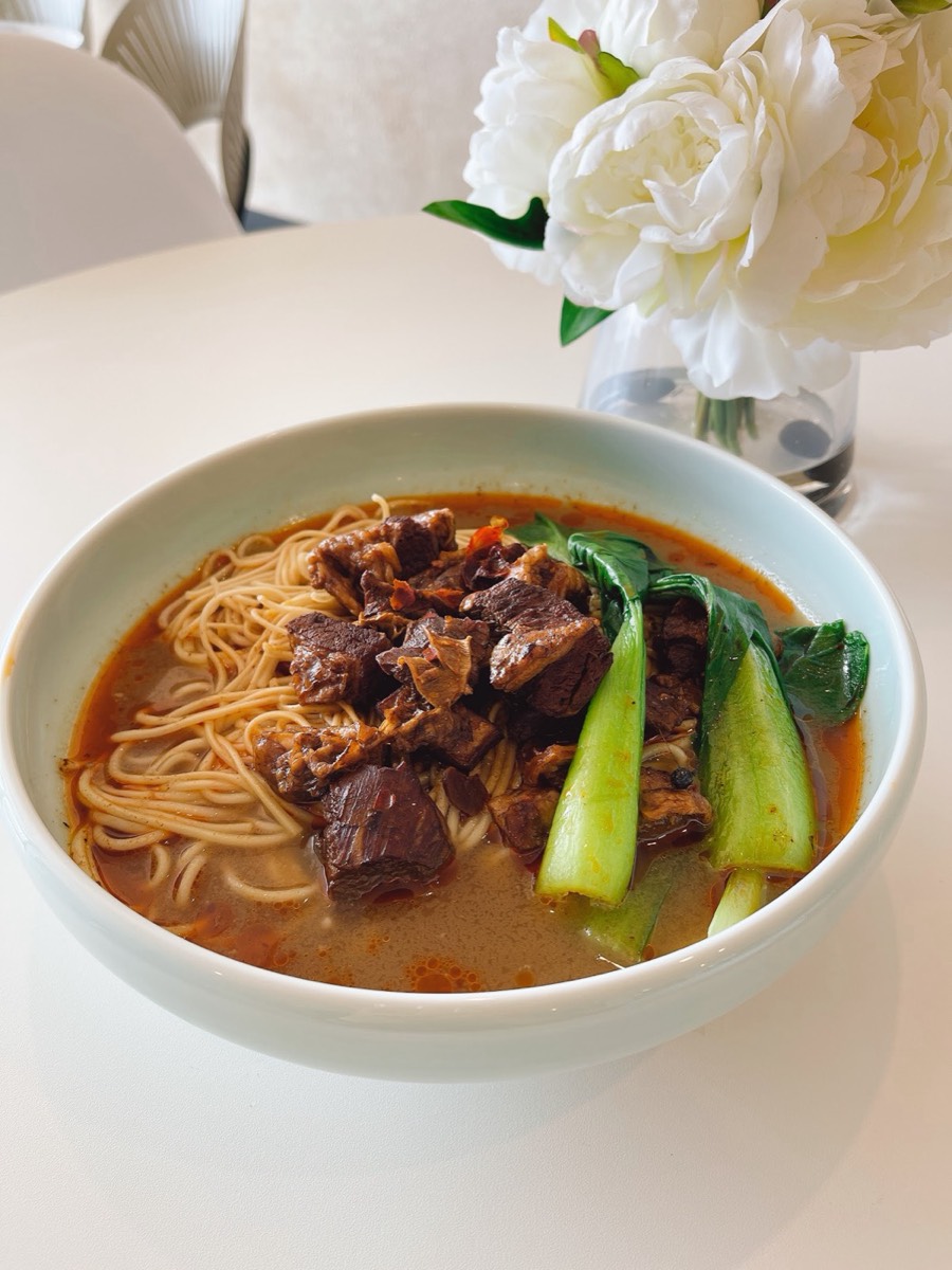 A white bowl of braised beef noodle soup with chunks of beef and bok choy, with white peonies on the table behind