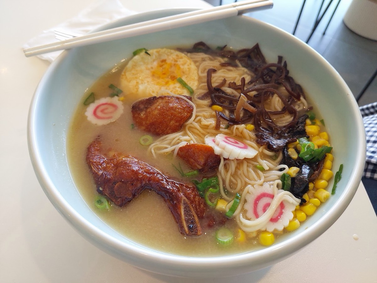A bowl of pale-broth ramen with a roast chicken drumstick, a soft egg, corn, black fungus and a pink-swirled fish cake