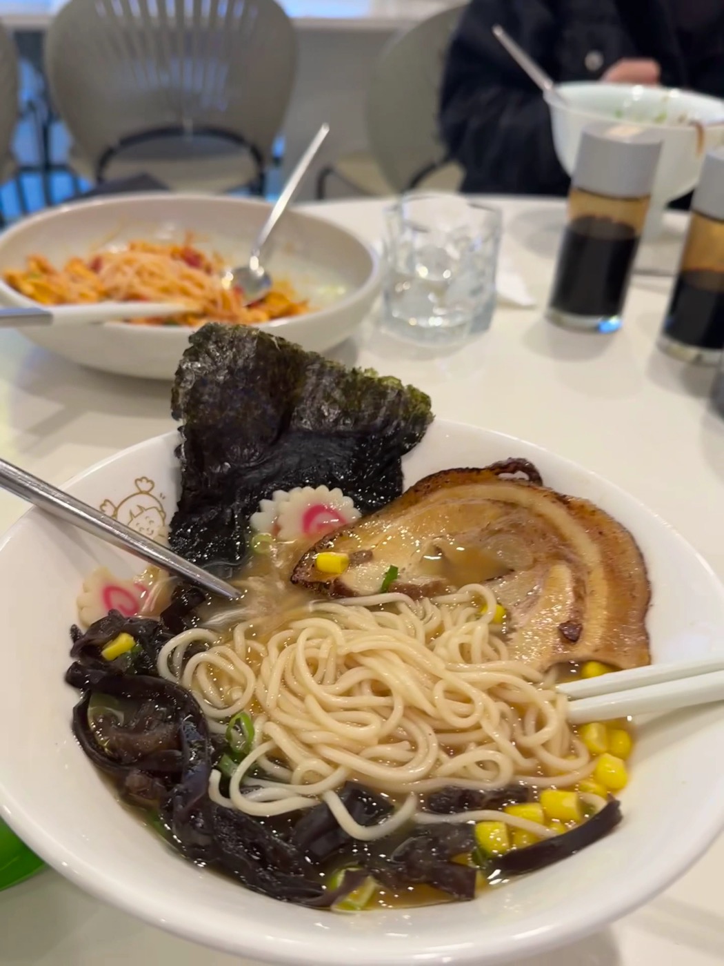 A bowl of ramen with a slice of roast pork, a sheet of nori, corn, black fungus and a pink-swirled fish cake