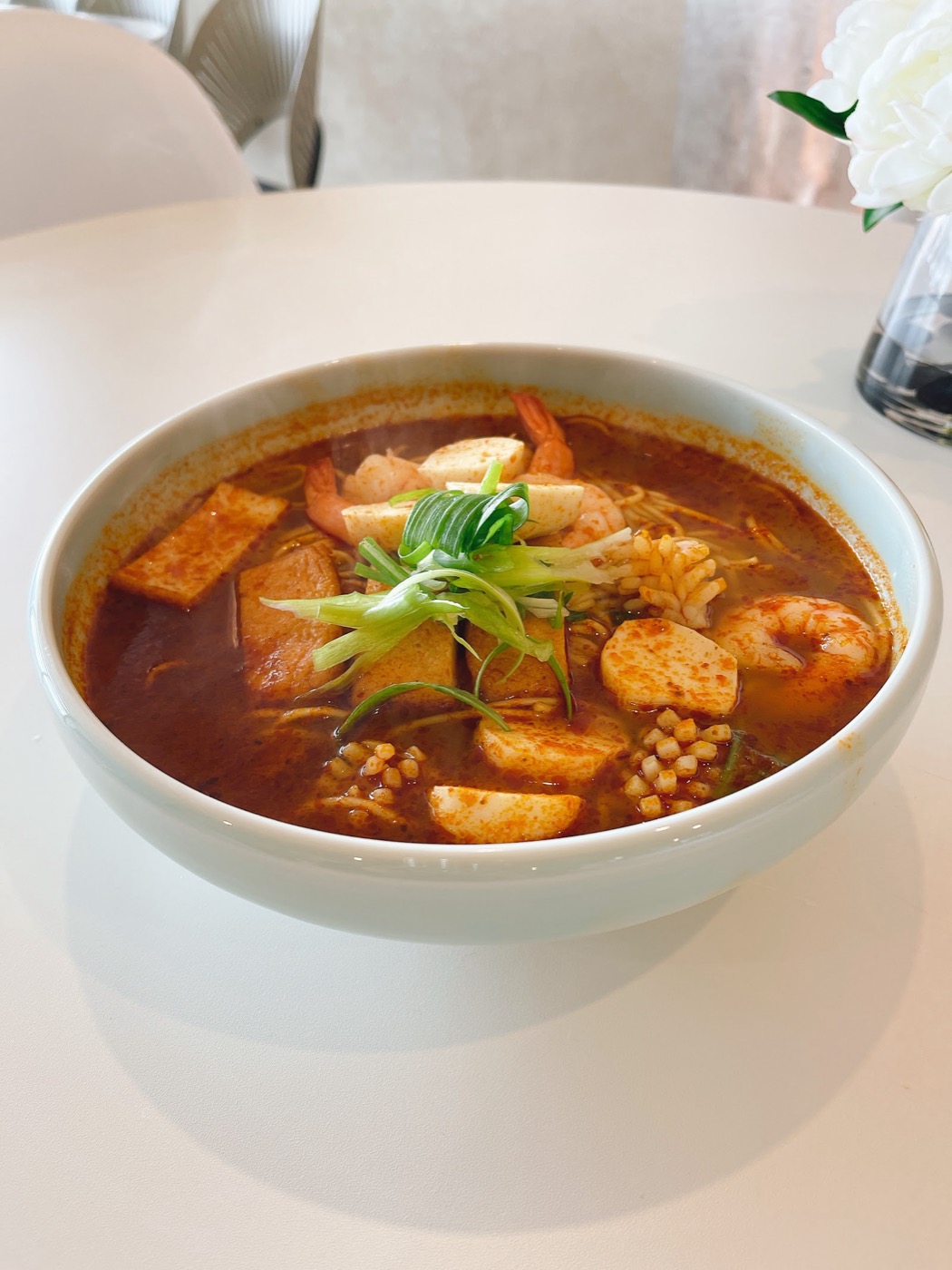 A white bowl of red-orange curry soup with prawns, squid, tofu and fish balls, topped with spring onion