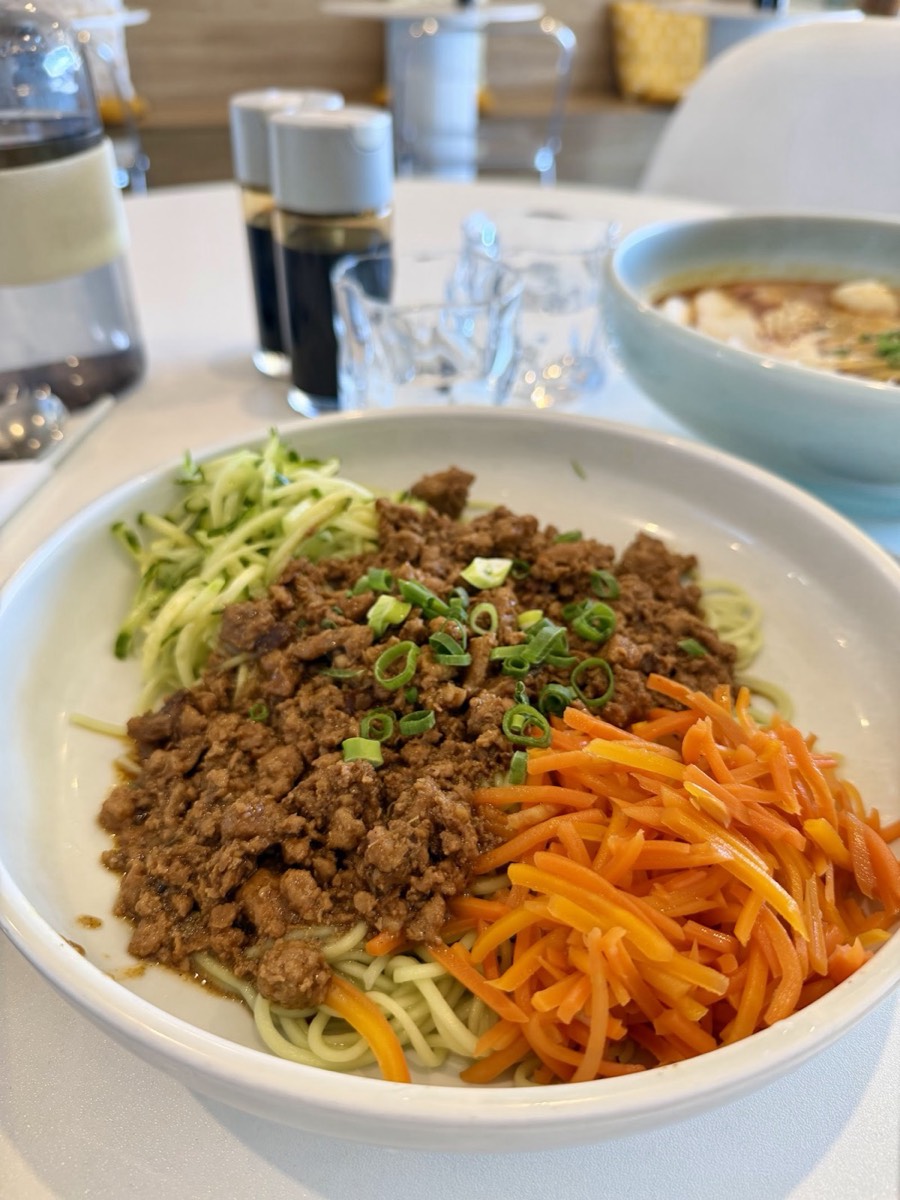 A white bowl of dry noodles topped with minced meat sauce, shredded cucumber and carrot